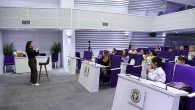 Konferans salonunda, mor koltuklarda oturan katılımcılara işaret dili öğreten bir kadın eğitmenin ayakta durarak ders anlatıyor.