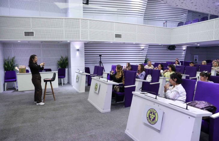 Konferans salonunda, mor koltuklarda oturan katılımcılara işaret dili öğreten bir kadın eğitmenin ayakta durarak ders anlatıyor.