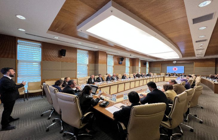 Görselde, büyük bir toplantı salonunda uzun bir masa etrafında toplanmış çok sayıda kişi görülüyor. Katılımcılar resmi kıyafetler içinde, toplantı sırasında notlar alıyor ve konuşmaları dinliyor. Salon modern bir tasarıma sahip; duvarda Türkiye Büyük Millet Meclisi logosu bulunan bir ekran yer alıyor.