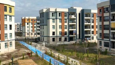 Yeni yapılmış modern apartman binalarının yer aldığı bir site görülüyor. Ortada mavi bir yürüyüş veya bisiklet yolu ve çevresinde genç ağaçlar ve yeşil alanlar bulunuyor.