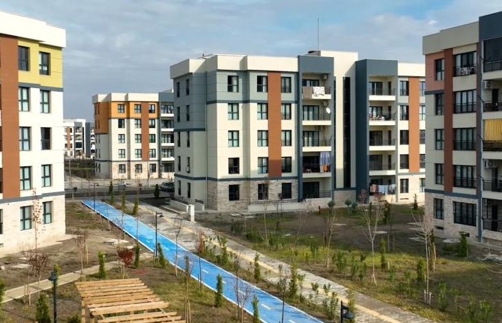 Yeni yapılmış modern apartman binalarının yer aldığı bir site görülüyor. Ortada mavi bir yürüyüş veya bisiklet yolu ve çevresinde genç ağaçlar ve yeşil alanlar bulunuyor.