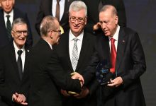 Bir ödül töreni sahnesi görünüyor. Ön planda takım elbiseli iki kişi tokalaşırken, sağda Cumhurbaşkanı Recep Tayyip Erdoğan elinde cam bir ödül plaketi tutuyor. Arkalarında yine takım elbiseli birkaç kişi daha ayakta duruyor ve sahne ışıkları tören havasını güçlendiriyor.