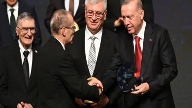 Bir ödül töreni sahnesi görünüyor. Ön planda takım elbiseli iki kişi tokalaşırken, sağda Cumhurbaşkanı Recep Tayyip Erdoğan elinde cam bir ödül plaketi tutuyor. Arkalarında yine takım elbiseli birkaç kişi daha ayakta duruyor ve sahne ışıkları tören havasını güçlendiriyor.