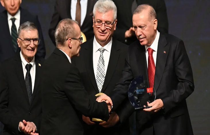 Bir ödül töreni sahnesi görünüyor. Ön planda takım elbiseli iki kişi tokalaşırken, sağda Cumhurbaşkanı Recep Tayyip Erdoğan elinde cam bir ödül plaketi tutuyor. Arkalarında yine takım elbiseli birkaç kişi daha ayakta duruyor ve sahne ışıkları tören havasını güçlendiriyor.