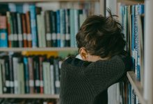 Bir kütüphanede, bir çocuk başını kollarına dayamış şekilde raflara yaslanmış duruyor; üzgün veya yorgun görünüyor.