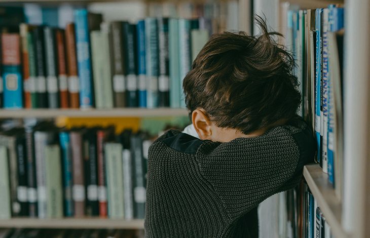 Bir kütüphanede, bir çocuk başını kollarına dayamış şekilde raflara yaslanmış duruyor; üzgün veya yorgun görünüyor.