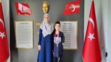 Bir kadın ve bir çocuk, arkalarında Atatürk büstü, Türk bayrakları ve İstiklal Marşı ile Gençliğe Hitabe panolarının bulunduğu bir duvar önünde poz veriyor. Çocuğun boynunda madalyalar asılı.