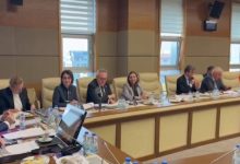 Bir toplantı salonunda uzun bir masanın etrafında oturan kişiler görülüyor. Önlerinde belgeler, dosyalar, mikrofonlar ve su şişeleri var. Katılımcılar not alıyor, konuşuyor ya da gündemi takip ediyor. Büyük pencerelerden gün ışığı geliyor. Ortam resmi, düzenli ve komisyon toplantısı havasında.