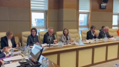 Bir toplantı salonunda uzun bir masanın etrafında oturan kişiler görülüyor. Önlerinde belgeler, dosyalar, mikrofonlar ve su şişeleri var. Katılımcılar not alıyor, konuşuyor ya da gündemi takip ediyor. Büyük pencerelerden gün ışığı geliyor. Ortam resmi, düzenli ve komisyon toplantısı havasında.