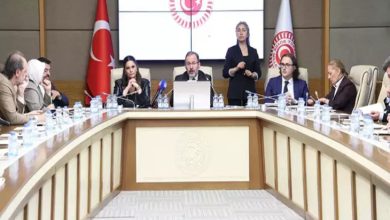 Görselde TBMM’de yapılan bir komisyon toplantısı görülüyor. Ortada mikrofonun başında konuşan bir kişi, etrafında ise masa boyunca oturmuş milletvekilleri ve katılımcılar yer alıyor. Arkada Türk bayrağı ve Türkiye Büyük Millet Meclisi amblemi bulunuyor. Toplantı resmî ve ciddi bir atmosferde gerçekleşiyor.