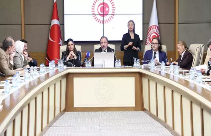 Görselde TBMM’de yapılan bir komisyon toplantısı görülüyor. Ortada mikrofonun başında konuşan bir kişi, etrafında ise masa boyunca oturmuş milletvekilleri ve katılımcılar yer alıyor. Arkada Türk bayrağı ve Türkiye Büyük Millet Meclisi amblemi bulunuyor. Toplantı resmî ve ciddi bir atmosferde gerçekleşiyor.