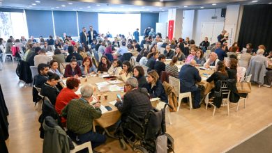 Geniş bir salonda çok sayıda kişi yuvarlak masalarda toplanmış halde çalıştay düzeninde oturuyor. Masalarda belgeler, notlar ve içecekler var; arka planda Türk bayrağı görülüyor.