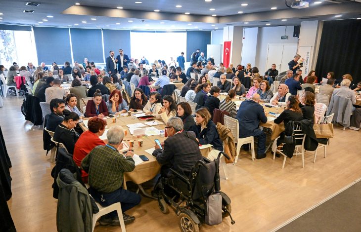 Geniş bir salonda çok sayıda kişi yuvarlak masalarda toplanmış halde çalıştay düzeninde oturuyor. Masalarda belgeler, notlar ve içecekler var; arka planda Türk bayrağı görülüyor.