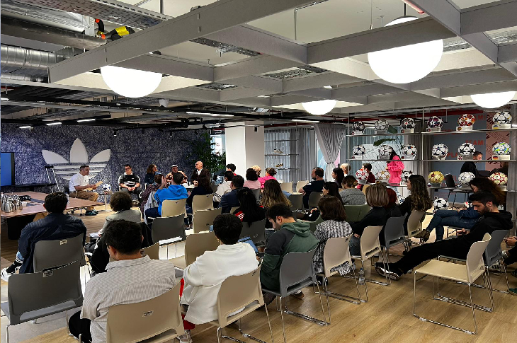 Görselde bir etkinlik salonunda düzenlenen söyleşi ya da panel oturumu görülüyor. Ön tarafta konuşmacılar sahnede otururken, karşılarında çok sayıda katılımcı sıralar halinde etkinliği izliyor. Salon modern bir tasarıma sahip; sol duvarda büyük bir Adidas logosu, sağ tarafta ise çeşitli futbol toplarının sergilendiği raflar yer alıyor. Ortam aydınlık ve düzenli, katılımın yoğun olduğu bir etkinlik atmosferi hakim.