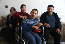 Bir ev ortamında, tekerlekli sandalyedeki bir erkek ön planda oturuyor. Yanında duran genç bir kadın ona destek oluyor ve gülümseyerek omzuna elini koyuyor. Arkada yaşlı bir kadın ve erkek, kanepeye oturmuş şekilde yer alıyorlar. Sıcak ve samimi bir aile atmosferi görülüyor.