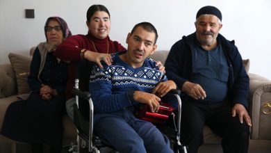 Bir ev ortamında, tekerlekli sandalyedeki bir erkek ön planda oturuyor. Yanında duran genç bir kadın ona destek oluyor ve gülümseyerek omzuna elini koyuyor. Arkada yaşlı bir kadın ve erkek, kanepeye oturmuş şekilde yer alıyorlar. Sıcak ve samimi bir aile atmosferi görülüyor.