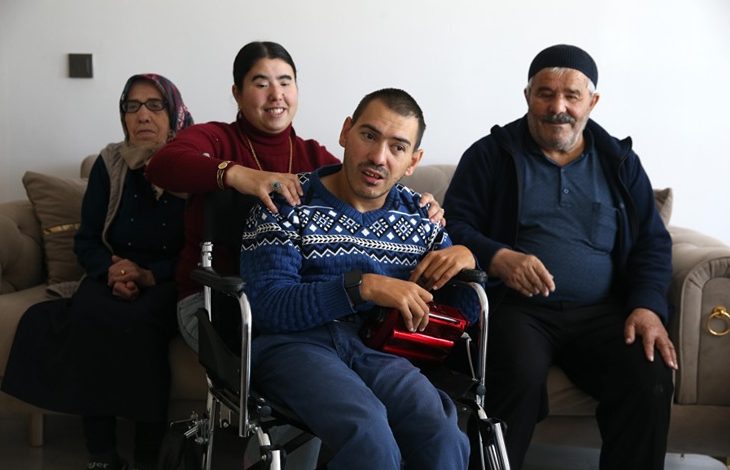 Bir ev ortamında, tekerlekli sandalyedeki bir erkek ön planda oturuyor. Yanında duran genç bir kadın ona destek oluyor ve gülümseyerek omzuna elini koyuyor. Arkada yaşlı bir kadın ve erkek, kanepeye oturmuş şekilde yer alıyorlar. Sıcak ve samimi bir aile atmosferi görülüyor.