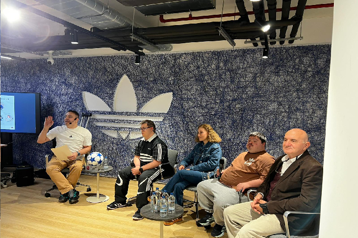 Görselde bir panel ya da sohbet etkinliği görülüyor. Beş kişi yan yana oturmuş; konuşmacılardan biri mikrofonla dinleyicilere hitap ediyor. Arkada büyük bir Adidas logosu bulunan modern bir iç mekân var. Masalarda su şişeleri duruyor ve ortam aydınlık, samimi bir etkileşim havası taşıyor.