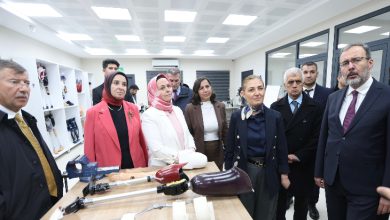 Aydınlık bir laboratuvar ortamında, bir grup kişi ortopedik ortez ve protezlerin sergilendiği bir çalışma masası etrafında toplanmış. Masada çeşitli protez bacak parçaları ve ekipmanlar duruyor. Grup dikkatle anlatılanları dinlerken ciddi ve ilgili bir atmosfer var. Arkada raflarda başka ortopedik cihazlar görülüyor.