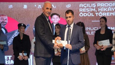 Görselde bir ödül töreni sahnesi yer alıyor. Önde, takım elbise giymiş iki erkek bulunuyor; biri diğerine ödül plaketini takdim ediyor. Arkada sahne dekorunda “Açılış Programı – Resim, Hikâye Ödül Töreni” yazısı görülüyor. Arka planda birkaç kadın ve erkek ödül alan katılımcılar sahnede yer almakta, ellerinde sertifika veya ödül plaketleri tutuyorlar.