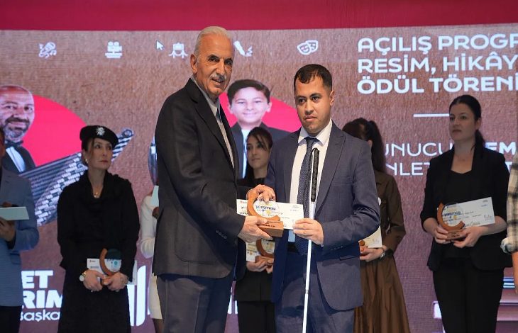 Görselde bir ödül töreni sahnesi yer alıyor. Önde, takım elbise giymiş iki erkek bulunuyor; biri diğerine ödül plaketini takdim ediyor. Arkada sahne dekorunda “Açılış Programı – Resim, Hikâye Ödül Töreni” yazısı görülüyor. Arka planda birkaç kadın ve erkek ödül alan katılımcılar sahnede yer almakta, ellerinde sertifika veya ödül plaketleri tutuyorlar.