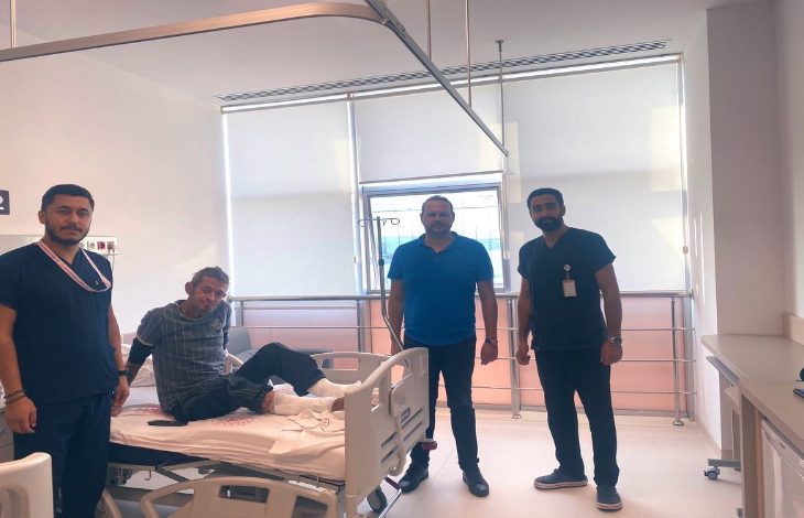 Aydınlık bir hastane odasında dört kişi görülüyor. Ortada, hastane yatağında oturan bir hasta var; ayakları bandajlı. Yanında üç sağlık çalışanı ya da görevli ayakta duruyor. Odanın büyük pencerelerinden gün ışığı süzülüyor, ortam temiz ve sakin bir tedavi alanı hissi veriyor.