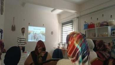 Bir sınıf ortamında, başörtülü kadınlardan oluşan bir grup sandalyelerde oturmuş eğitmene doğru bakıyor. Eğitmen, projeksiyonla yansıtılan “Fiziksel erişim engelleri” başlıklı bir görsel eşliğinde sunum yapıyor. Odada raflarda çantalar ve el işi ürünler de görülüyor.