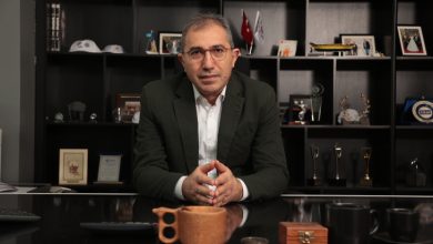 Fotoğrafta bir ofiste, İDO Genel Müdürü Dr. Murat Orhan, takım elbiseli, gözlüklü bir takarak başında oturuyor. Arkasında ödüller ve kurumsal objelerle dolu raflar var. Masada ahşap bir fincan, kupalar ve küçük eşyalar duruyor. Genel hava profesyonel, ciddi ve düzenli.