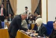 Bir toplantı salonunda sıralı masalarda oturan kalabalık bir grup görünüyor. İnsanlar evraklara, bilgisayarlara ve notlara yoğunlaşmış hâlde. Önde, takım elbiseli bir erkek eğilerek bir kadınla konuşuyor ya da bir şey gösteriyor. Ortam ciddi, bürokratik ve yoğun çalışma havasında.