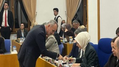 Bir toplantı salonunda sıralı masalarda oturan kalabalık bir grup görünüyor. İnsanlar evraklara, bilgisayarlara ve notlara yoğunlaşmış hâlde. Önde, takım elbiseli bir erkek eğilerek bir kadınla konuşuyor ya da bir şey gösteriyor. Ortam ciddi, bürokratik ve yoğun çalışma havasında.