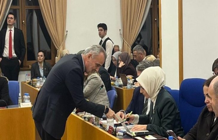 Bir toplantı salonunda sıralı masalarda oturan kalabalık bir grup görünüyor. İnsanlar evraklara, bilgisayarlara ve notlara yoğunlaşmış hâlde. Önde, takım elbiseli bir erkek eğilerek bir kadınla konuşuyor ya da bir şey gösteriyor. Ortam ciddi, bürokratik ve yoğun çalışma havasında.