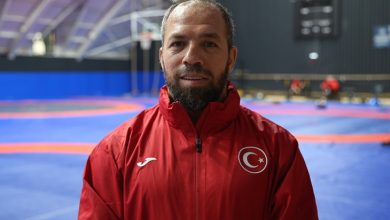 Kırmızı milli eşofman üstü giymiş işitme engelli sporcu Oğuz Dönder, minderlerin bulunduğu kapalı bir antrenman salonunda kameraya bakıyor. Arka planda mavi–turuncu güreş minderleri ve antrenman yapan kişiler seçiliyor.