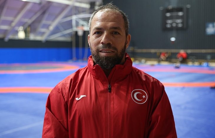 Kırmızı milli eşofman üstü giymiş işitme engelli sporcu Oğuz Dönder, minderlerin bulunduğu kapalı bir antrenman salonunda kameraya bakıyor. Arka planda mavi–turuncu güreş minderleri ve antrenman yapan kişiler seçiliyor.