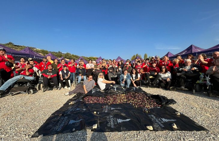 Açık havada düzenlenen etkinlikte, engelli bireyler ve yakınları topluca poz veriyor. Önlerinde zeytinlerle dolu örtüler, arkalarında mor çadırlar ve masmavi bir gökyüzü görülüyor. Herkes neşeli ve dayanışma içinde.