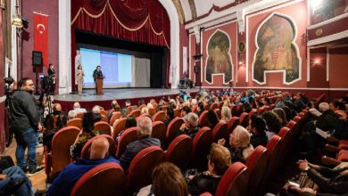 Tarihi bir tiyatro salonunda, kırmızı koltuklara oturmuş kalabalık bir izleyici grubunun karşısında, sahnede bir sunucu ve yanında işaret diliyle çeviri yapan bir kişiyle bir sunum gerçekleştiriliyor.
