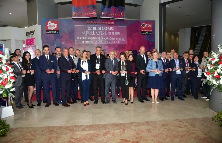 Geniş bir kalabalığın katıldığı resmi bir açılış töreni görülüyor. Uzun bir kurdele boyunca yan yana dizilmiş katılımcılar, makaslarıyla toplu bir kurdele kesimine hazırlanıyor. Arkada büyük bir afiş, bilimsel kongre ve sergi etkinliğinin adını taşıyor. Herkes şık giyimli, ortamda tören havası ve heyecanlı bir kalabalık enerjisi var.