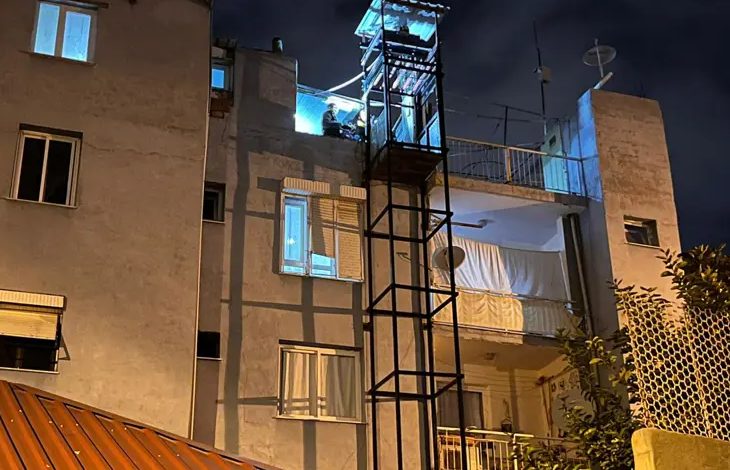 Fotoğrafta, çok katlı bir apartmanın dış cephesine sonradan eklenmiş bir engelli asansörü görülüyor. Gece çekilmiş görüntüde asansör kabini en üst kata yakın bir noktada, mavi ışıkla aydınlanmış şekilde duruyor. Binanın cephesi eski bir yapıyı andırıyor; açık balkonlar, perdeyle kapatılmış alanlar ve farklı tipte pencereler dikkat çekiyor. Asansörün metal iskeleti dar ve oldukça yüksek; bu da kullanımı sırasında ciddi riskler oluşturabilecek bir görünüm veriyor.