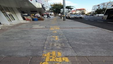 Bir kaldırım üzerinde yer alan sarı hissedilebilir yürüme yolu, yer yer aşınmış ve bozulmuş halde görülüyor. Yolun ortasında bir aydınlatma direği bulunuyor, yanında araçlar ve dükkânlar yer alıyor.