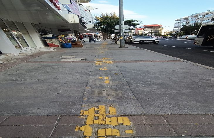 Bir kaldırım üzerinde yer alan sarı hissedilebilir yürüme yolu, yer yer aşınmış ve bozulmuş halde görülüyor. Yolun ortasında bir aydınlatma direği bulunuyor, yanında araçlar ve dükkânlar yer alıyor.