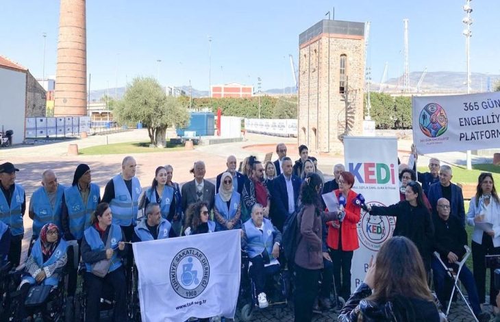 Görselde açık havada düzenlenmiş bir basın açıklaması görülüyor. Ön tarafta tekerlekli sandalyedeki bireyler ve yanlarında ayakta duran katılımcılar bulunuyor. Bir grup, üzerinde Türkiye Sakatlar Derneği amblemi olan büyük bir beyaz bayrak tutuyor. Diğer tarafta “KEDİ” logosu ve “365 Gün Engellilik Platformu” yazılı pankartlar yer alıyor. Katılımcılar bir araya gelmiş, dayanışma ve farkındalık temasında bir açıklama yapıyor gibi görünüyor.