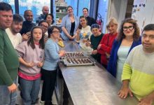 Görselde, bir mutfakta tezgâhın etrafında toplanmış bir grup kişi görülüyor. Tezgâh üzerinde tepsiyle hazırlanmış çikolata ya da kurabiye benzeri yiyecekler var. Katılımcılar gülümseyerek poz veriyor. Ortam, mutfak becerileri eğitimi ya da atölye çalışması havasında.