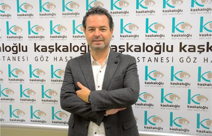 Görselde bir göz hastanesinin tanıtım panosu önünde duran Op. Dr. Sedat Selim görülüyor. Üzerinde koyu renk bir ceket ve beyaz gömlek var. Kollarını önde birleştirerek poz vermiş. Arka planda tekrarlayan “Kaşkaloğlu Göz Hastanesi” yazıları ve göz logosu yer alıyor. Ortam profesyonel ve sağlık kurumuna ait bir tanıtım havası taşıyor.