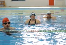 Görselde bir yüzme havuzunda üç genç yüzücü yan yana görülüyor. Her biri bone ve yüzücü gözlüğü takmış, suyun içinde antrenman arasında dinleniyor gibi duruyorlar. Arka planda kulvar ipleri ve kapalı havuz ortamının duvarları seçiliyor. Atmosfer sakin, odaklı bir antrenman havası veriyor.