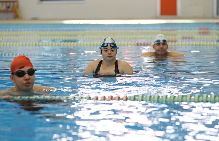 Görselde bir yüzme havuzunda üç genç yüzücü yan yana görülüyor. Her biri bone ve yüzücü gözlüğü takmış, suyun içinde antrenman arasında dinleniyor gibi duruyorlar. Arka planda kulvar ipleri ve kapalı havuz ortamının duvarları seçiliyor. Atmosfer sakin, odaklı bir antrenman havası veriyor.