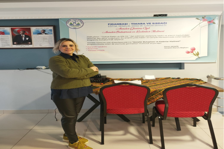 Görselde, bir kadın ayakta durup kameraya gülümseyerek poz veriyor. Arkasında dernek etkinliğine ait büyük bir afiş, duvarda ise resmi bir portre bulunuyor. Önde kırmızı sandalyeli bir masa ve üzerinde örtü serili bir düzenleme yer alıyor. Ortam bir toplantı ya da dernek salonunu andırıyor.