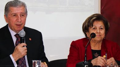 Solda takım elbiseli Köksal Toptan, elinde mikrofonla masada oturmuş konuşuyor. Sağda kırmızı ceketli Saime Toptan da oturuyor.