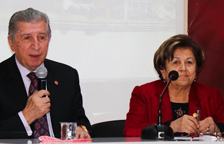 Solda takım elbiseli Köksal Toptan, elinde mikrofonla masada oturmuş konuşuyor. Sağda kırmızı ceketli Saime Toptan da oturuyor.