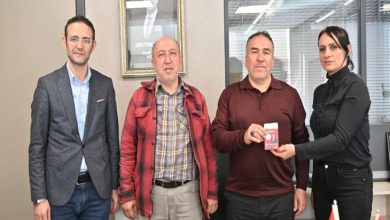 Görselde dört kişi yan yana ayakta poz veriyor. Sağdaki kadın, yanında duran erkeğe kırmızı bir kart ya da cihaz uzatıyor; erkek bunu elinde tutuyor. Ortam bir ofis gibi görünüyor; arkada cam bölmeler ve duvarda bir portre bulunuyor. Sol tarafta da iki erkek daha yer alıyor. Genel olarak resmi bir ziyaret veya teslim töreni havası var.