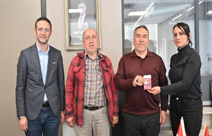 Görselde dört kişi yan yana ayakta poz veriyor. Sağdaki kadın, yanında duran erkeğe kırmızı bir kart ya da cihaz uzatıyor; erkek bunu elinde tutuyor. Ortam bir ofis gibi görünüyor; arkada cam bölmeler ve duvarda bir portre bulunuyor. Sol tarafta da iki erkek daha yer alıyor. Genel olarak resmi bir ziyaret veya teslim töreni havası var.