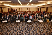 Görselde geniş bir konferans salonunda kalabalık bir katılımcı grubu görülüyor. Ön sırada resmi kıyafetli katılımcılar otururken, arkada salonu dolduran çok sayıda kişi dikkatle etkinliği takip ediyor. Ortam, bir konferans veya ödül törenine ait düzenli ve resmi bir atmosfer yansıtıyor.